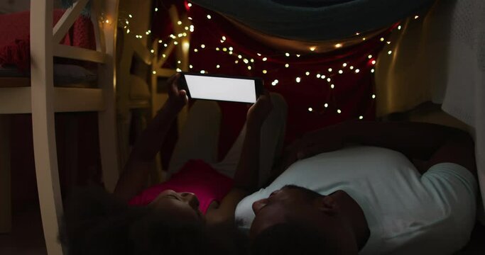 African american daughter and her father watching tablet lying in blanket tent with fairy lights