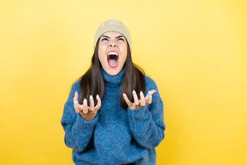 Young beautiful woman wearing blue casual sweater and wool hat crazy and mad shouting and yelling...