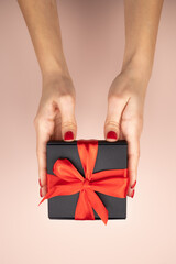 Female hands hold out wrapped black gift box with red ribbon on pink background.