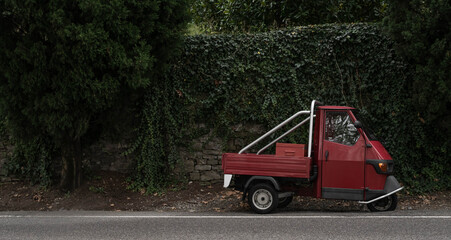 Red, little retro car or delivery truck on the road among green wall of plants. Typical red and green italian picture. in dark, dull colors.