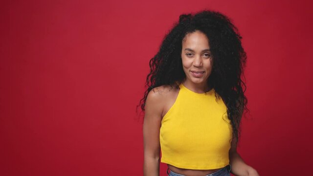 Cheerful african american young woman in yellow tank top posing isolated on red background studio. People lifestyle concept. Doing dab hip hop dance hands move gesture youth sign hiding covering face