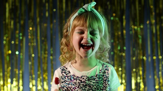 Futuristic hipster teen girl with lollipop candy looking at camera smiling, screaming, shouting in slow motion at disco party cyberpunk club. Child kid in neon lights standing by illuminated wall