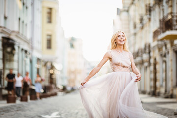 Fototapeta na wymiar Pretty smiling young woman with long blond hair in elegant flying light dress running along the street