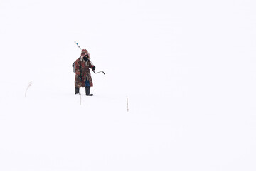 Fototapeta premium A lone fisherman in warm clothes and rubberized felt boots with an ice drill on his shoulder walks along a snow-covered river. Copy space.