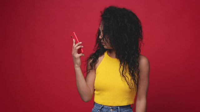 Displeased irritated african american woman in casual yellow tank top isolated on red color background studio. People lifestyle concept. Talking on mobile phone showing blah blah gesture ja jaja hands