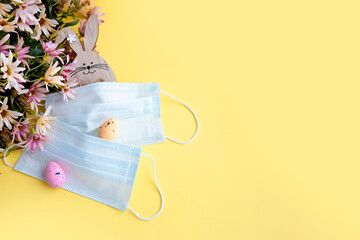 Medical protective face mask, flower, wooden bunny and colorful easter eggs isolated on yellow background. Concept of Easter holiday during pandemic coronavirus. Flat lay, top view, copy space.