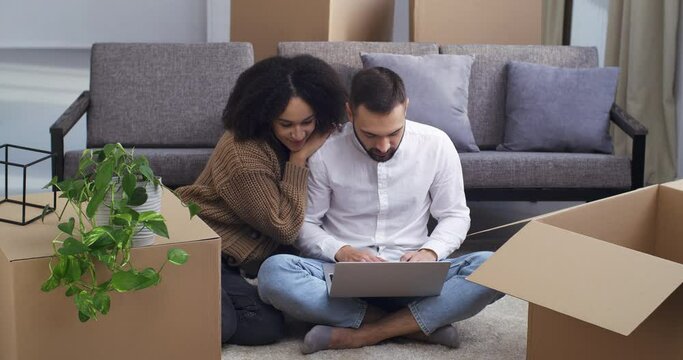 Mixed race couple multiethnic family african american woman and caucasian man sitting on floor of new house surrounded by boxes choosing goods online shopping online ordering freight service to move