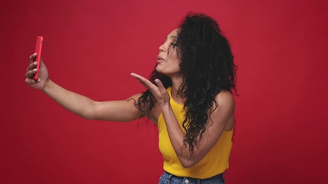 Smiling african american woman in yellow tank top isolated on red background studio. People lifestyle concept. Doing selfie shot on mobile phone showing victory sign thumb up blowing sending air kiss