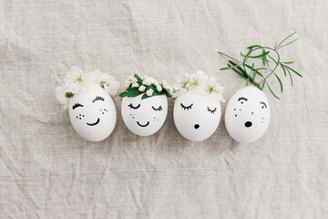 Natural eggs with drawn funny faces in cute floral crowns top view on linen fabric with petals
