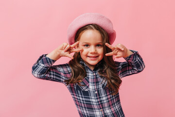 Charming little girl in pink beret and blue shirt shows peace sign on pink background