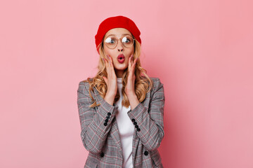 Attractive blonde girl in stylish outfit is looking at camera in surprise. Woman in red beret and gray jacket posing on pink background