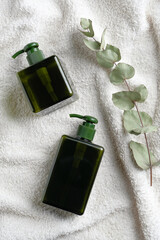 SPA skincare products design. Green glass pump cosmetic bottles and eucalyptus leaf on white towel. Flat lay, top view.
