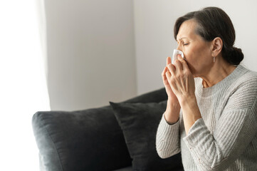 Sick senior woman has cold and flu, sneezes and has runny nose, wipes her nose with a paper tissue sitting on the couch at home. Viral disease concept