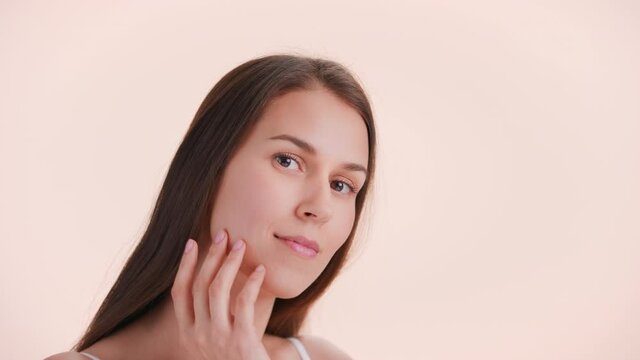 Beauty portrait of young brown-haired woman with perfect skin slowly touches her face sliding fingers from cheekbone down to the chin and looks at camera | Beauty products commercial concept