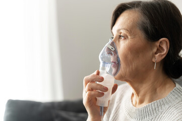 Side view a senior older woman is using inhaler for flu and cold treatment sitting on the sofa at home. Healthcare and medicine concept - obrazy, fototapety, plakaty