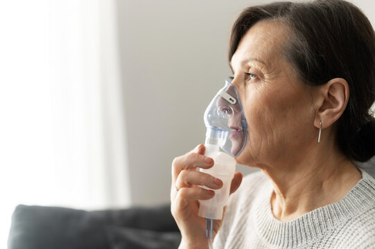 Side view a senior older woman is using inhaler for flu and cold treatment sitting on the sofa at home. Healthcare and medicine concept