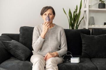 Senior woman using inhaler for flu and cold treatment sitting on the sofa at home. Healthcare and...