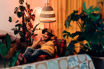 relaxed man with cowboy hat lying down on a lounge at home holding telephone and typing on the...