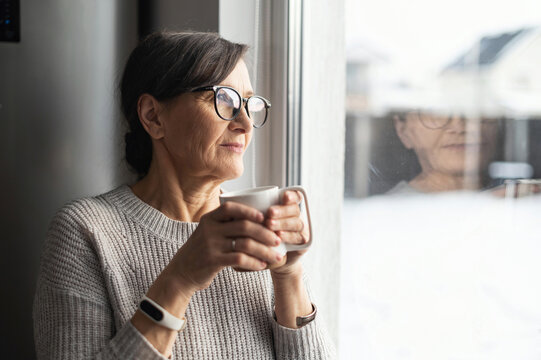 Close-up portrait of senior older woman wearing glasses enjoys morning coffee in the kitchen at home. A modern retirement lady daydreaming with a mug of hot drink looks through the window - Powered by Adobe