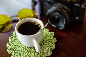 Xícara com café, câmera antiga e óculos com lentes amarelas sobre uma mesa de madeira.