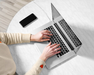 Fototapeta premium Close up of female hands on a laptop keyboard view from above. Young woman using a computer while sitting at an office table. Nearby lies a smartphone