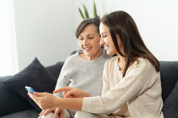 An adult daughter helps to mother figures out how to use a smartphone, two multigenerational women sitting on the couch and look at phone screen
