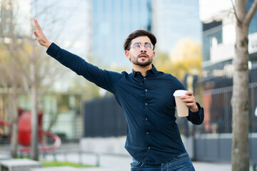 Young man holding a cup of coffee and calling a taxi outdoors.