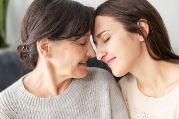 A senior mother and an adult daughter sitting on the comfortable couch face to face, a mom and a daughter touching foreheads with eyes closed. Family ties