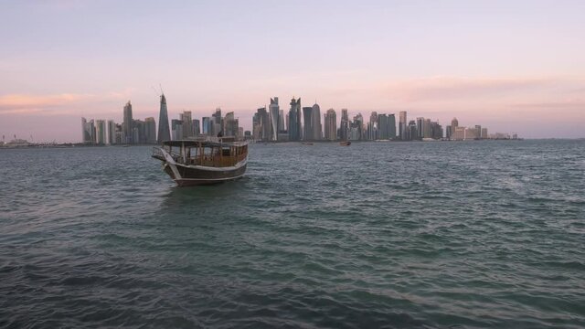 Doha skyline from the corniche promenade sunset zooming in shot showing dhow with Qatar flags in the Arabic gulf  in foreground and clouds in the sky in background
