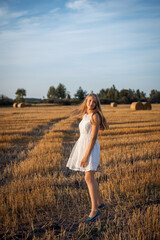 Photo of a blond girl wearing a white dress walking in the field of harvested rye. Attractive girl in the agricultural field in the evening