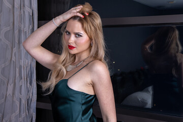 Fototapeta na wymiar An elegant attractive slim woman with long blonde hair and red lipstick wearing a long green evening dress, looking sexy and seductive near a large window
