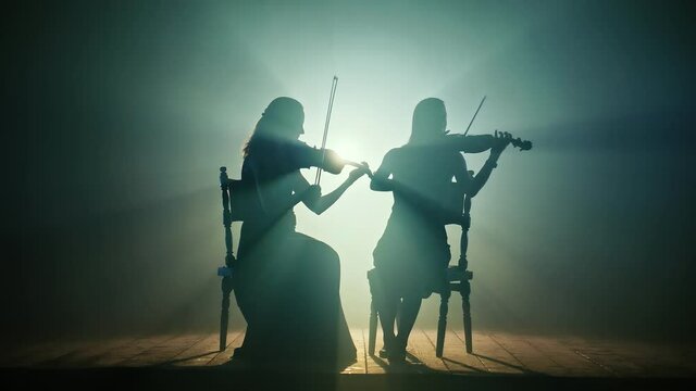 Silhouettes of Girls Violinists Professionally Playing Violins on the Large Stage of the Chamber Hall in Smoke on a Dark Background with Illumination.