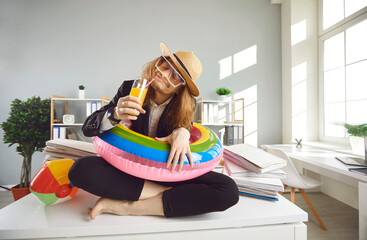 Young office worker in beach sunhat, sunglasses and inflatable swim ring sitting on desk, dreaming...