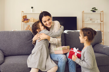 Happy young woman sitting on sofa at home on Mother's Day, hugging her children and thanking them...