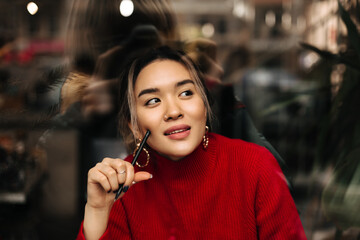 Beautiful brown-eyed woman in big gold earrings and red outfit posing thoughtfully with pen in her hands