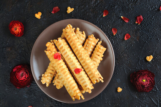 Golden Wafer Rolls On A Brown Plate With Red Petals And Flowers On A Black Background. View From Above