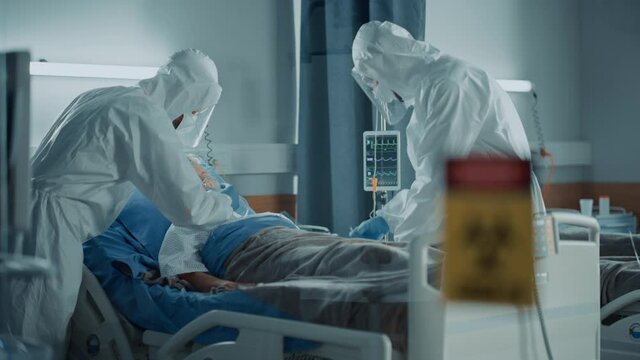 Hospital Coronavirus Emergency Department Ward: Doctors wearing Coveralls, Face Masks Save Life of a Senior Patient Lying in Bed, Put Oxygen Mask and Connect Iv Drip. Biohazard Sign on Door
