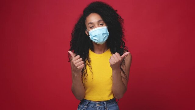 Funny african american woman in yellow tank top face mask safe from coronavirus virus covid-19 isolated on red background. People lifestyle concept. Point finger camera on you showing thumb up gesture