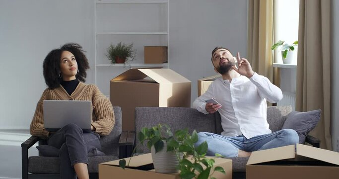 Multiethnic couple surrounded by cardboard boxes sitting on couch choosing new home looking for apartment using laptop and phone planning dreaming presenting ordering furniture parcels online in net