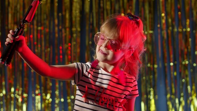 Fashion girl in futuristic night club with red neon lights making selfie call on mobile phone. Trendy stylish child kid blogger smiling cheerfully, recording video blog vlog on smartphone with tripod