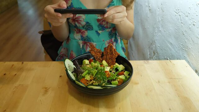 Girl take picture of avocado salad using smartphone, bowl with tasty food stand on wooden table. Close shot of freshly cooked meal at cosy cafe, late breakfast time at Spain