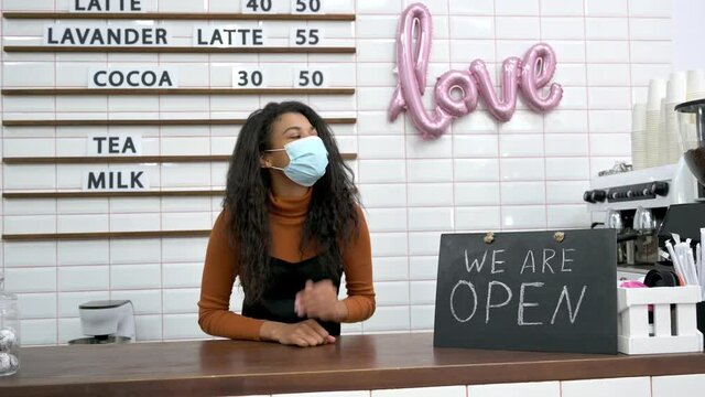 A friendly African American female barista, waitress or cafe bar owner wearing a medical mask, stands behind the bar counter next to a signboard WE ARE OPEN, waiting for visitors