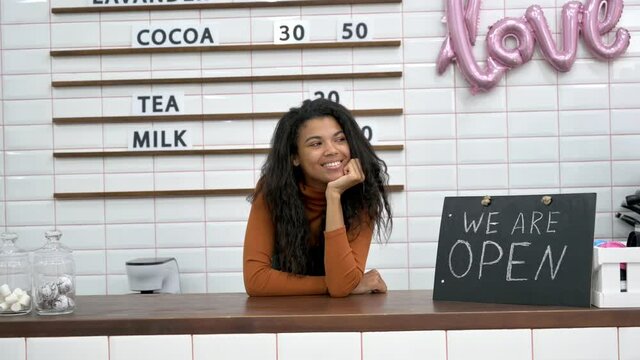 Happy African American female barista or waitress, owner of a coffee shop, stands near a signboard WE ARE OPEN behind the bar counter and friendly smiling