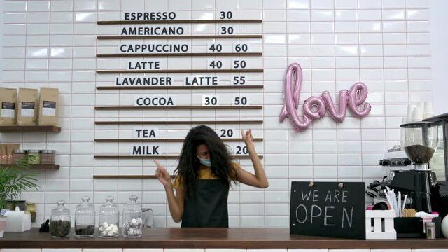 Happy African American curly girl, waitress, barista or coffee shop owner, in a medical mask, dances and rejoices at the opening of a coffee bar, there is an OPEN sign on the counter