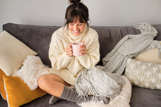 Happy woman having coffee in mug while sitting on sofa at home during winter