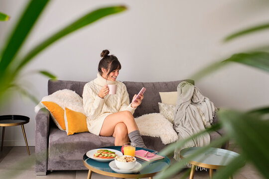 Smiling woman using smart phone while having coffee on sofa at home during winter