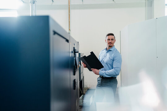 Male professional with file document looking away while standing by machine in factory