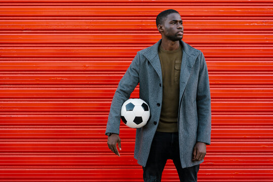 Young man with soccer ball looking away while standing against red wall