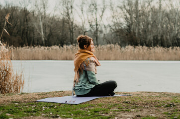 Fototapeta premium Back view of young woman in comfy clothes seated in meditation on the ground in a park, with a lake background. Concept: self care practices, movement, introspection