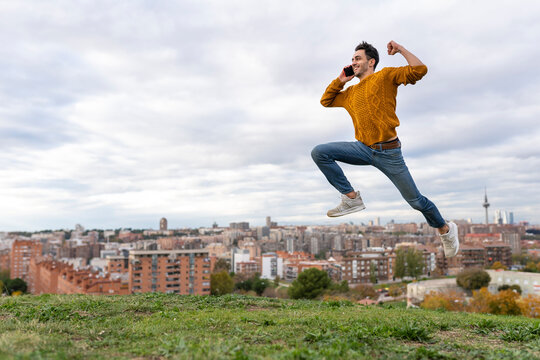Excited young man talking on mobile phone while jumping over hill against cityscape